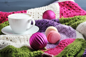 On a knitted multi-colored plaid white cup with cappuccino. Near two pastry macaroons. Two Christmas balls give the composition a Christmas mood. Close-up.
