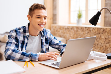 I feel happy. Handsome exuberant fair-haired boy working on his laptop and smiling while sitting at the table