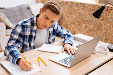 Learning lessons. Attractive serious fair-haired boy working on his laptop and writing in his notebook while sitting at the table