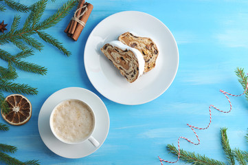 Several slices of Stollen with raisins and marzipan filling and a cup of coffee. Christmas composition is complemented by a spruce branch and a slice of orange. Blue wood background. View from above.