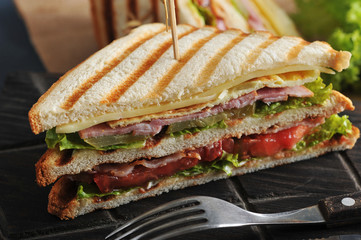 Half of a sandwich in the form of a triangle on a wooden board. In the background, the other half of the sandwich. There are cutlery nearby. Dark wooden background. Close-up. Macro photography.