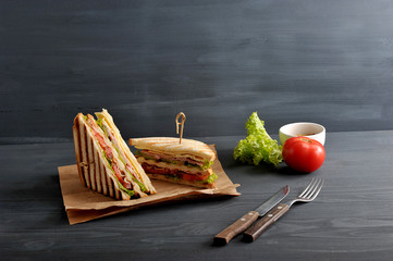 Two halves of the club sandwich on kraft paper. The filling of the sandwich consists of ham, cheese, tomato, bacon, sauce and fried egg. Nearby cutlery, mustard, vegetables. Dark background. Close-up.