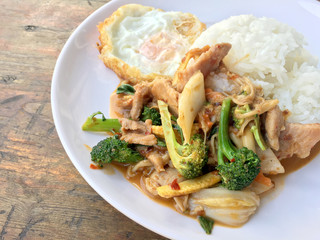 Asian style stir-fry meat slices with fresh vegetable with rice and fried eggs in thai style placed on white dish on wooden table.