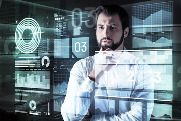 How interesting. Smart concentrated attentive programmer standing in front of a futuristic screen and looking at it while thoughtfully touching his beard