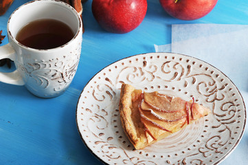 A piece of apple pie on a plate with a pattern. In the background a few red apples and a cup of tea. Blue wood background. Close-up. Macro photography.