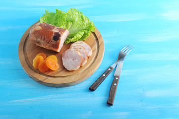 Meat roll with dried fruits from poultry meat on a wooden tray. Next to the roll, several cut slices and dried apricots. Blue wood background. View from above. Close-up. In the frame cutlery.