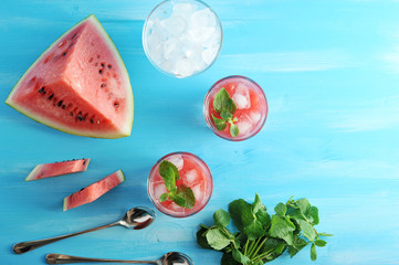 Two glasses of watermelon smoothies are decorated with fresh mint leaves. Nearby a few slices of watermelon, a bunch of fresh mint, a glass of ice and a quarter of a watermelon. View from above. 