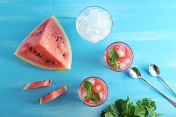 Two glasses with smuzzy from a water-melon. Near the glass with ice, a piece of watermelon, green mint and two teaspoons. Blue wood background. View from above. Close-up.