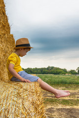 Naklejka na ściany i meble Little nice boy dressed in a yellow shirt and denim shorts sits on a haystack in a summer day. Harvesting.