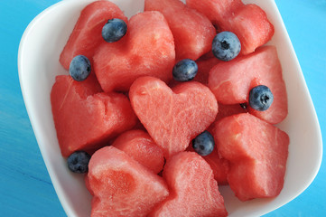 In a white plate pieces of a watermelon in the form of heart and a berry of a blueberry. Blue wood background. View from above. Close-up. Macro photography.