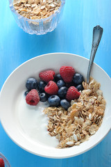 Muesli, yogurt, raspberries and blueberries. Next to the glass cup with muesli. There's a spoon in the plate. Blue wood background. View from above. Close-up. Vertical orientation of the frame.