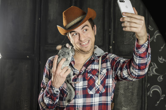 Portrait of a Cowboy. dark hair, jeans , plaid shirt. he holds the chinchilla animal . photographing self on smartphone