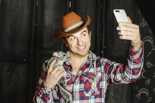 Portrait of a Cowboy. dark hair, jeans , plaid shirt. he holds the chinchilla animal . photographing self on smartphone