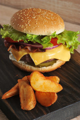 Hamburger and potatoes are rustic on a wooden board. The hamburger stuffing includes chopped meat cutlet, fried bacon, lettuce leaves, cheese, tomatoes, sauce. Vertical orientation of the frame. 