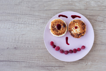 cheesecakes on a pink plate spread out in the form of a pensive smiley decorated with raspberries and jam on a wooden table