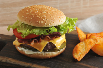 Hamburger and potatoes are rustic on a wooden board. The hamburger stuffing includes chopped meat cutlet, fried bacon, lettuce leaves, cheese, tomatoes, sauce. 