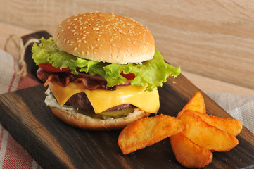 Appetizing cheeseburger and potatoes are rustic on a wooden board. In the cheeseburger filling, Cheddar cheese and fried bacon. Close-up.