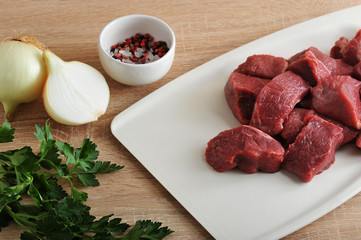 Pieces of beef on a white tray. Next to onions, parsley and a cup of pepper and salt. A set for cooking minced meat. Close-up. Light wooden background.