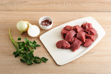 A set for cooking minced meat. Pieces of beef on a white tray. Next to onions, parsley and a cup of pepper and salt. Close-up. Light wooden background.