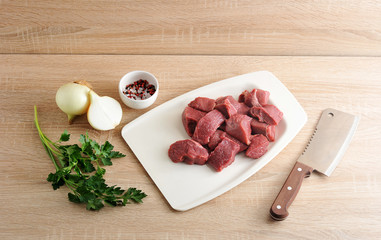A set for cooking minced meat. Pieces of beef on a white tray, onion, parsley and a cup of pepper and salt. Next a knife for cutting meat. Light wooden background.