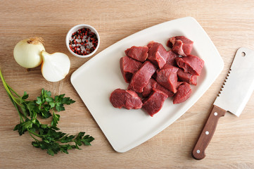 A set for cooking minced meat. Pieces of beef on a white tray, onion, parsley and a cup of pepper and salt. Next a knife for cutting meat. View from above. Close-up. Light wooden background.