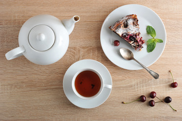 Portion of chocolate brownies with cherries on a plate. A cup of tea and a teapot. Fresh cherries are on the table. Serving for one person. View from above. Close-up.