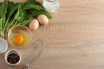 Spinach greens, cream in a glass jug, chicken eggs, a plate with a raw egg without a shell, a set of spices (salt, pepper), whisk for whipping. Set for filling open cake pie. Wooden beige background.