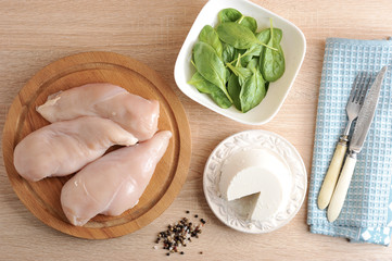 Chicken breast on a wooden tray. Ricotta cheese on a white plate and spinach greens. Set for cooking. Cutlery: fork and knife  on a napkin.  Beige wood background. VIew from above.