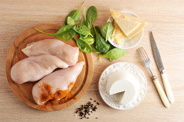 Chicken breast on a wooden tray. Ricotta cheese, spinach greens and Parmesan cheese. Set for cooking. Cutlery: fork and knife with white handles.  VIew from above.  Beige wood background.