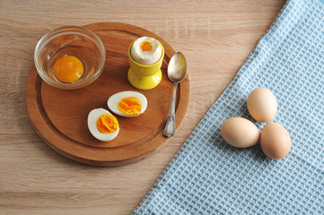 Raw chicken eggs on a cloth napkin. Next on a wooden tray are two halves of an hard-boiled egg, an soft-boiled egg and a glass bowl with a raw egg without a shell. 