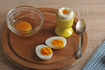 On a wooden tray are two halves of an hard-boiled eggs, an soft-boiled egg and a glass bowl with broken raw egg. Next teaspoon. Wooden light background. Closeup.