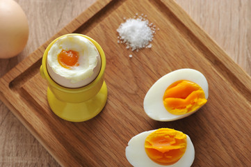On a wooden tray, an soft-boiled egg, salt and two halves of an hard-boiled egg. Wooden light background. View from above. Close-up. 