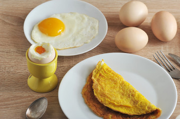 Omelette from chicken eggs, fried eggs, soft-boiled eggs. Cutlery. In the distance, hard-boiled eggs. Light wooden background.