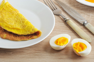 Omelette from chicken eggs on a white plate. Next two halves of  an hard-boiled eggs. Cutlery: fork and knife. Closeup.