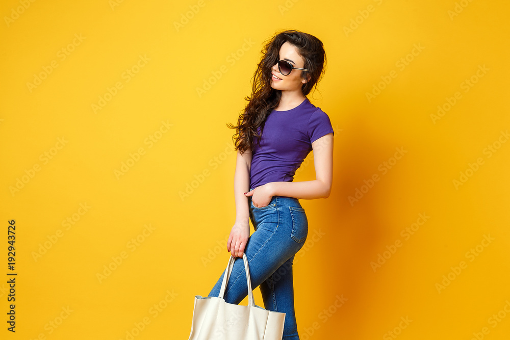 Wall mural Beautiful young woman in sunglasses, purple shirt, blue jeans posing with bag on the wonderful yellow background