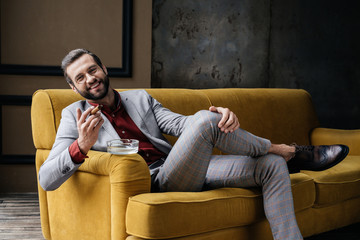 handsome laughing man with cigar and ashtray sitting on sofa  in loft interior