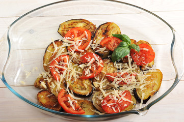 Vegetables baked with cheese and seasonings. In the aubergine, tomatoes, basil, olive oil, cheese and seasonings. A dish for baking from glass. Light wooden background.
