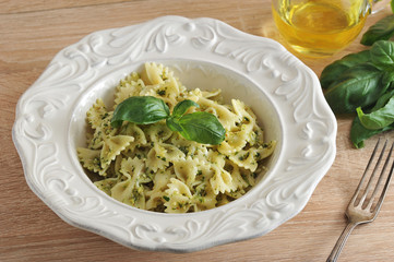 Pasta farfalle with pesto sauce in a white plate. The dish is decorated with a leaf of fresh basil. Next to the plate is a fork, a jug of olive oil and a bunch of fresh basil. Close-up.