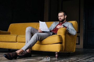 Fototapeta premium stylish man reading newspaper and sitting on couch, ashtray with cigar on floor