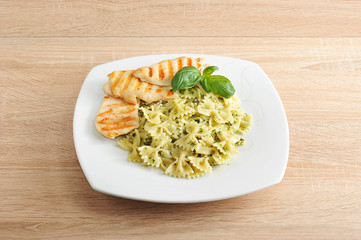 Pasta farfalle with pesto sauce on a white square plate. Next to the chicken breast, cooked on the grill.  The dish is decorated with a leaf of fresh basil. Close-up. Light wooden background.