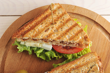 Sandwich with tuna from white toasted bread. Filling of tuna, eggs, tomato, green salad. The sandwich is on a wooden round tray. There are several olives nearby. View from above. Close-up.