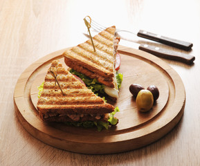 Sandwich with tuna from white toasted bread. Filling of tuna, eggs, tomato, green salad. The sandwich is on a wooden round tray. There are several olives nearby. View from above. Macro photography.