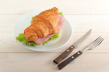 Croissant with a stuffing. As stuffing ham, green salad, tomato and cheese. There are cutlery nearby. Light wooden background. Close-up.