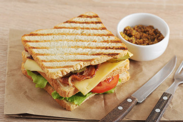 Classic club sandwich with a filling of chicken breast, bacon, green salad, cheese, tomato and pickled cucumbers. Next cutlery and a cup of mustard. Close-up.