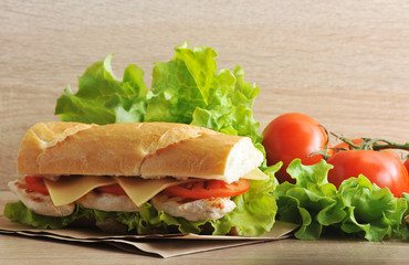 Sandwich of baguette on kraft paper. The filling of the sandwich consists of the leaves of a green salad, a chicken breast cooked on a grill, slices of cheese and a tomato. 