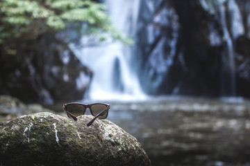Sunglasses left on the stone in front off small waterfall and green tree as background.