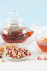 A teapot with black tea and a cup of tea. Glass ware. Next to the plate are dried rosebuds, used for brewing tea. Light background. Close-up. Vertical orientation of the frame
