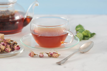 A teapot with black tea and a cup of tea. Glass ware. Next to the plate are dried rosebuds, used...