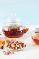 A teapot with black tea and a cup of tea. Glass ware. Next to the plate are dried rosebuds used for brewing tea. Light background. Close-up. Vertical orientation of the frame.