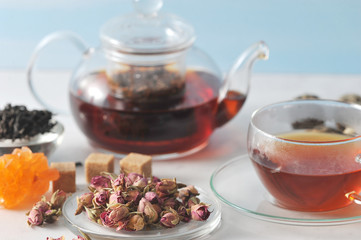 A teapot with black tea and a cup of tea. Glass ware. Next to the plate are dried rosebuds, used for brewing tea. In the frame, crystal sugar and tea leaves. Light background. Close-up. 